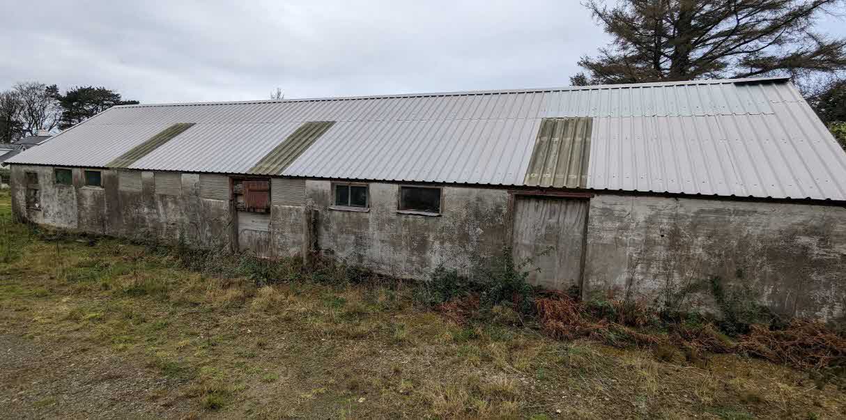 A photograph of a long, single-story agricultural barn with a corrugated metal roof and weathered concrete walls situated in a grassy field.