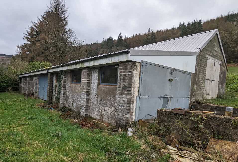 A photograph showing a long, single-story brick and concrete agricultural building with large metal doors in a rural setting.