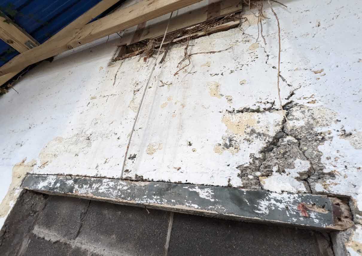 A close-up photograph showing the dilapidated condition of a white rendered wall with peeling plaster and exposed concrete, likely part of a barn structure.