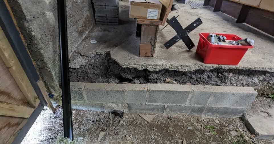 A photograph showing the interior of a building under construction, featuring a newly laid blockwork wall and a broken concrete floor with tools and materials.