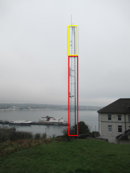 A photograph of a tall telecommunications mast with colored boxes highlighting sections of the structure, situated on a hill overlooking the sea.