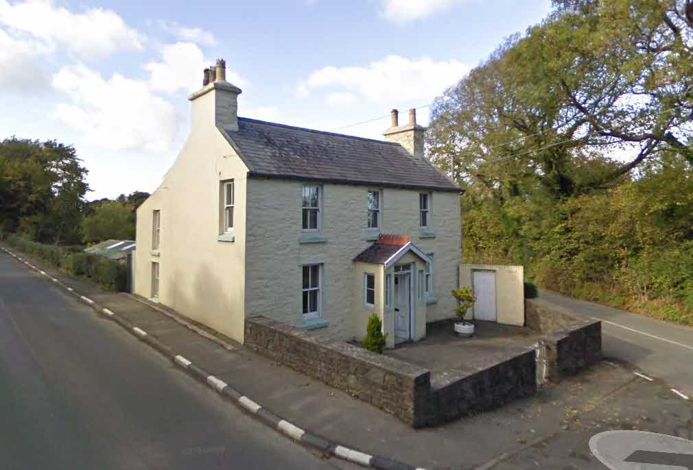 A photograph of a two-story detached stone house painted cream, situated on a street corner with a low stone wall and garden area.