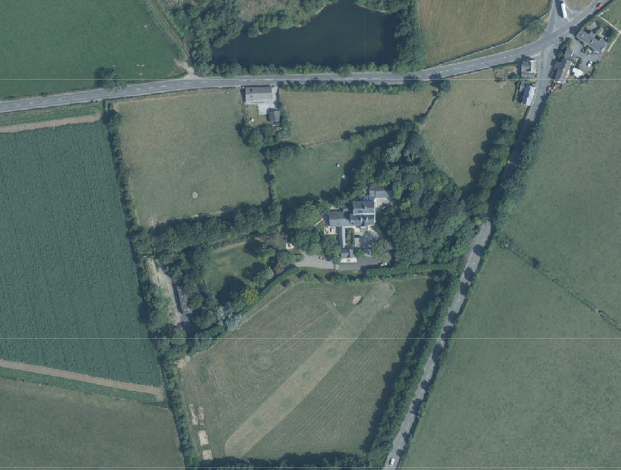 An aerial view of a rural property showing a house surrounded by trees and fields, with a road running along the top edge.