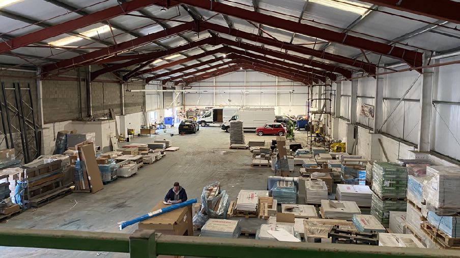Interior photograph of a large industrial warehouse unit featuring red steel roof trusses, stacks of building materials on pallets, and vehicles parked inside.