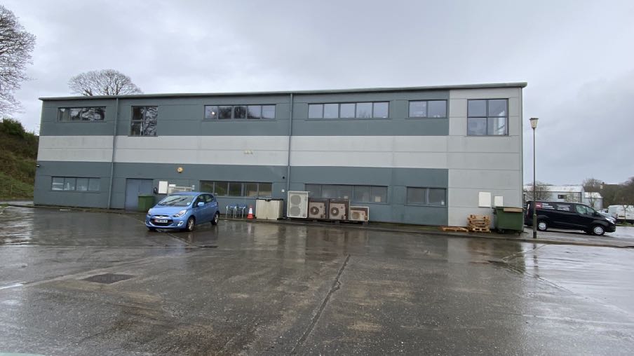A photograph showing the exterior of a two-story grey and white industrial or commercial building with a large paved parking area in front.