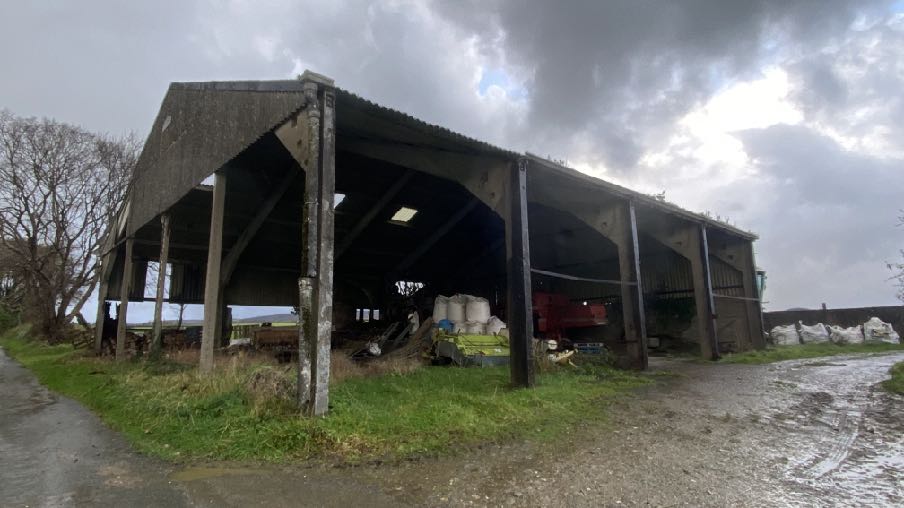 A photograph of a large, open-sided agricultural barn with concrete supports and a corrugated roof, containing farm machinery.