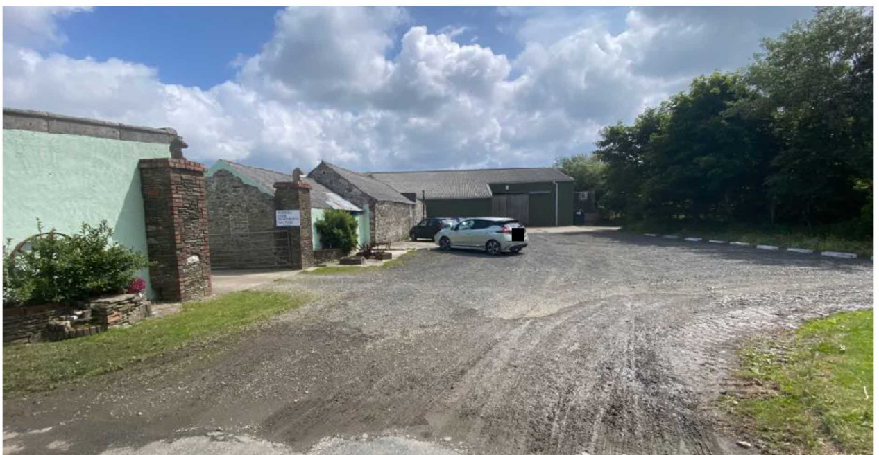A photograph of the existing rural site showing a gravel driveway, stone agricultural buildings, and a green building with a brick gate pillar.