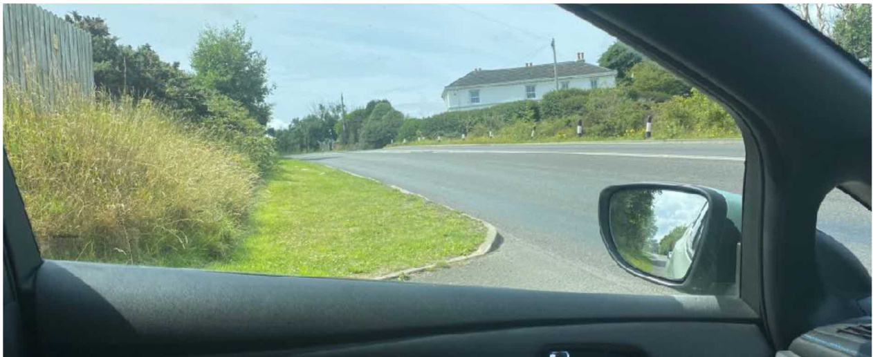 A photograph taken from inside a vehicle looking out at a rural road with a white house on a hill. Tall grass and a wooden fence are visible on the roadside.