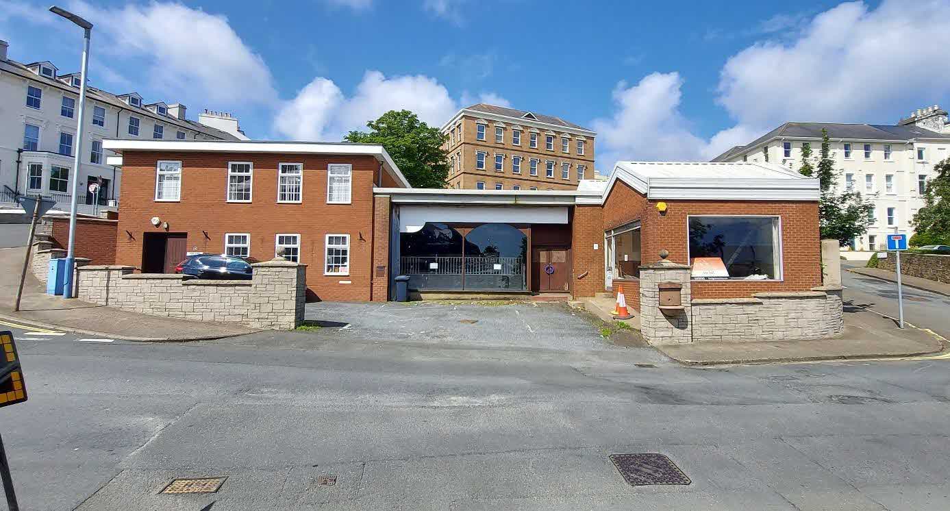 A street-level photograph showing a red brick building complex with a gated entrance and adjacent single-story structures, set against a backdrop of other residential buildings.
