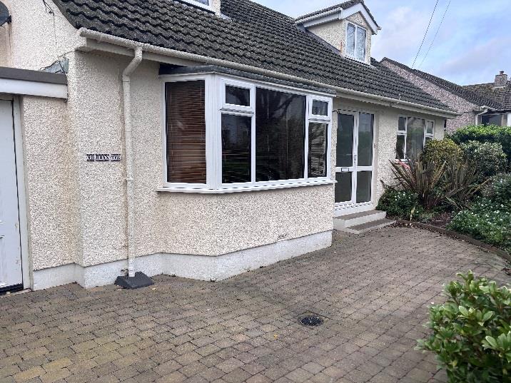 A photograph showing the front exterior of a detached bungalow with pebbledash walls, featuring a large bay window and a paved driveway. A garage door is visible on the left side of the building.