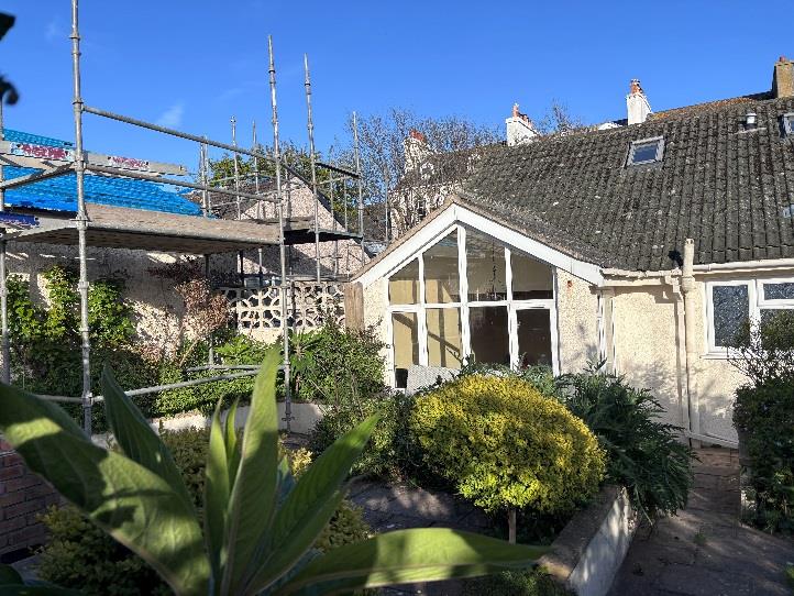 A photograph showing a residential property with scaffolding erected around a garage structure on the left and a house with a conservatory on the right.