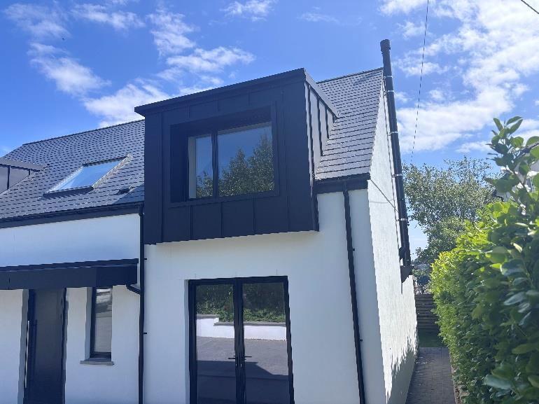 A photograph showing the rear elevation of a white house featuring a modern two-storey extension and a black dormer structure on the roof.
