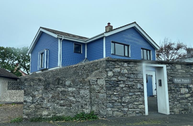 A photograph showing a blue two-story house with white trim situated behind a high, rough stone boundary wall with a white gate.