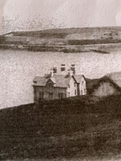 A grainy, black and white historical photograph showing a detached house situated on a hillside with a body of water in the background.