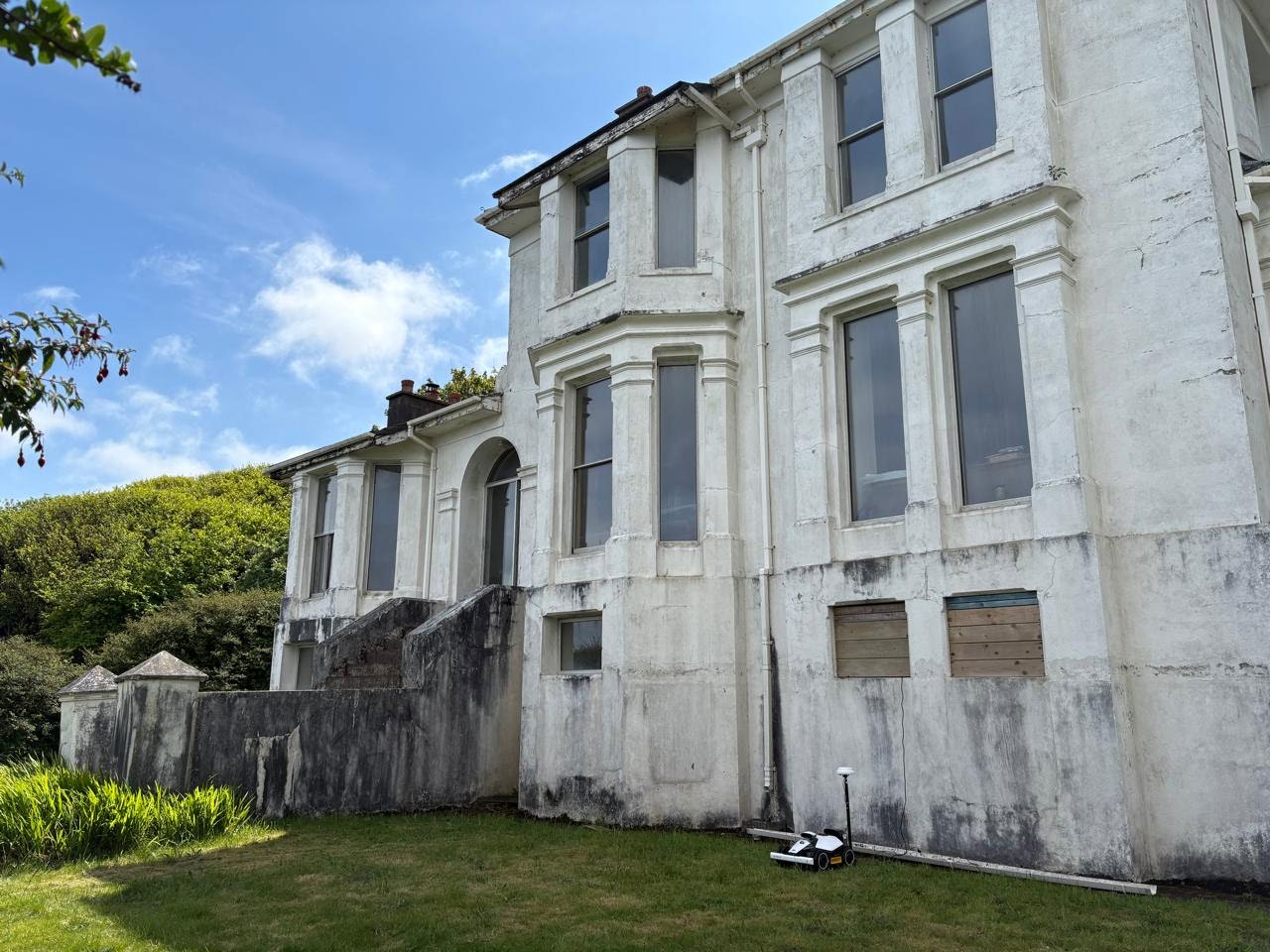 A photograph showing the side elevation of a large, white-rendered detached building with boarded ground floor windows and a surveying instrument on the lawn.
