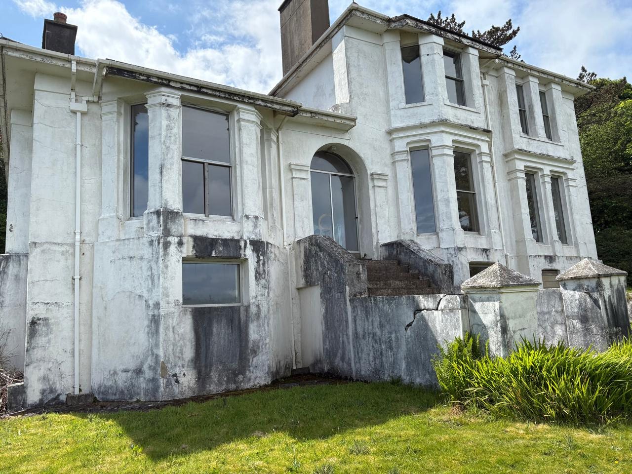 A photograph showing the exterior elevation of a large, white, multi-story building with weathered stucco and large sash windows. The building features a prominent chimney and is set behind a low stone wall with green...