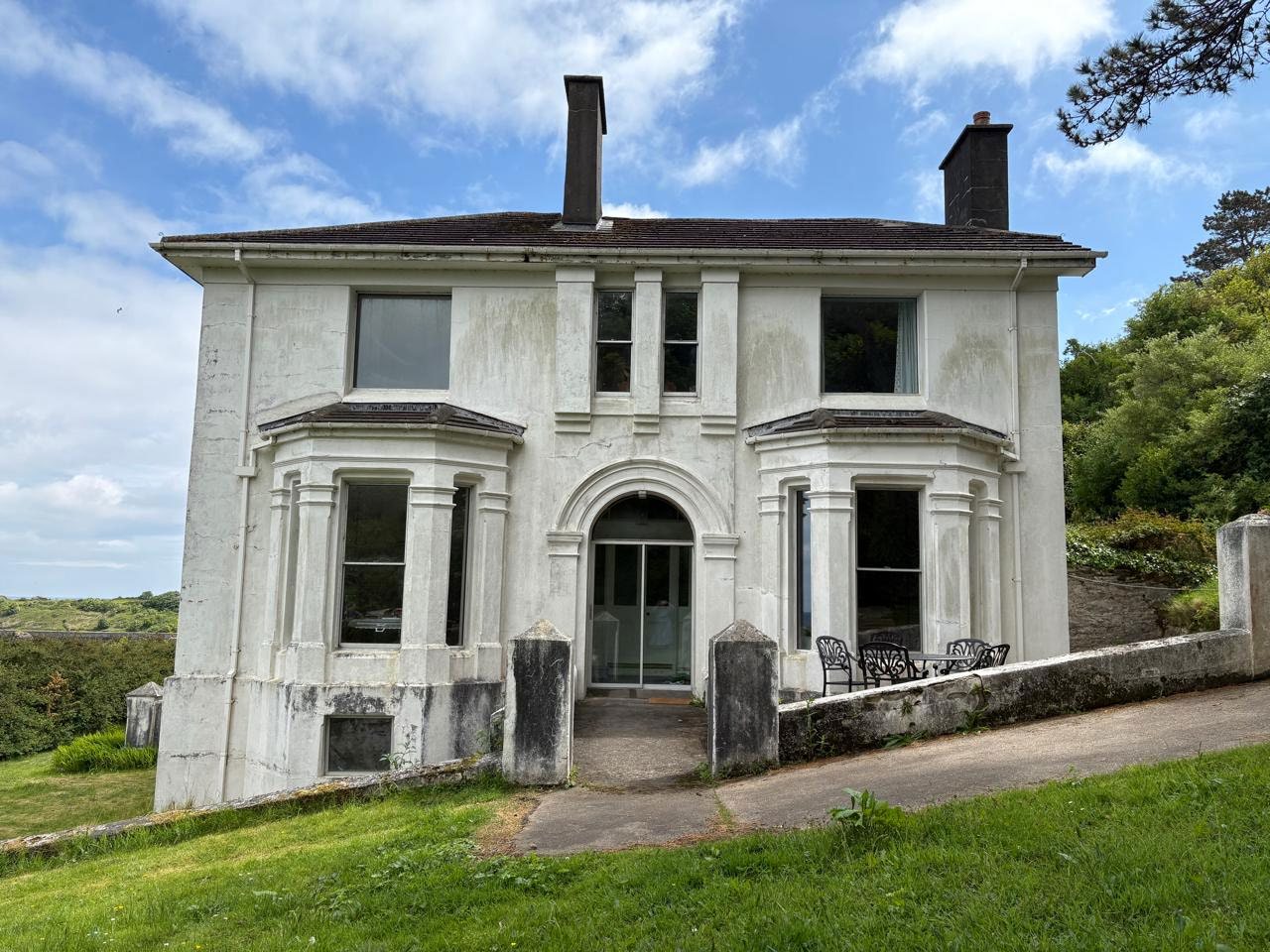 A photograph of a white, two-story detached house featuring bay windows and chimneys, situated on a grassy slope.