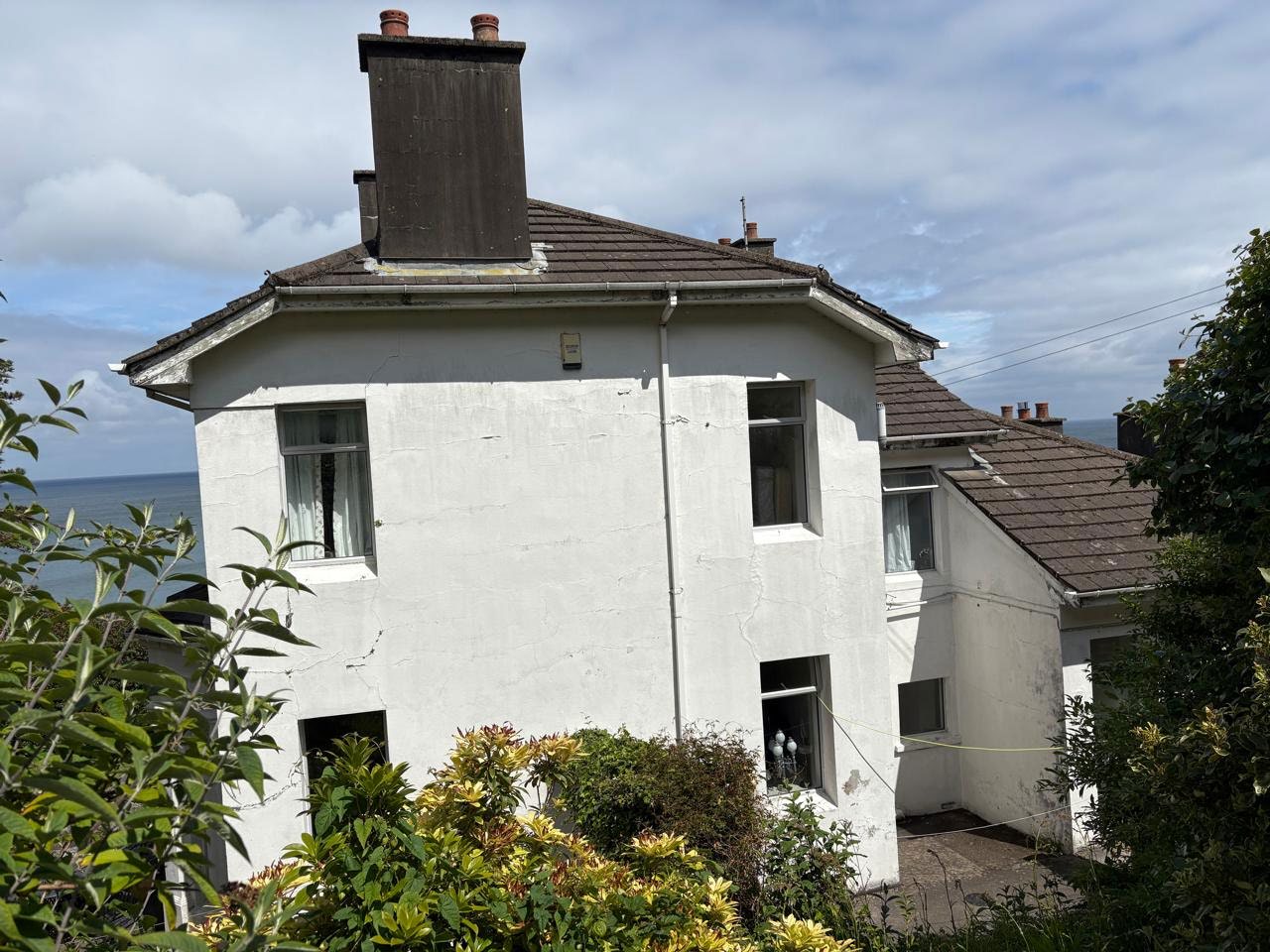 A white two-story house with a tiled roof and prominent chimney stack, situated near the coast with the sea visible in the background.