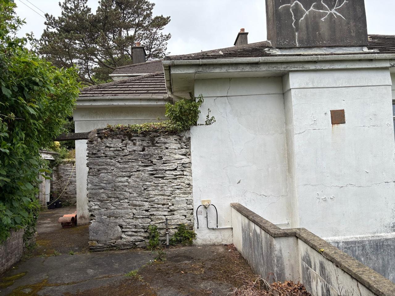A photograph showing the exterior of a residential property featuring a white rendered wall, an attached dry stone wall section, and a tiled roof with a chimney stack.