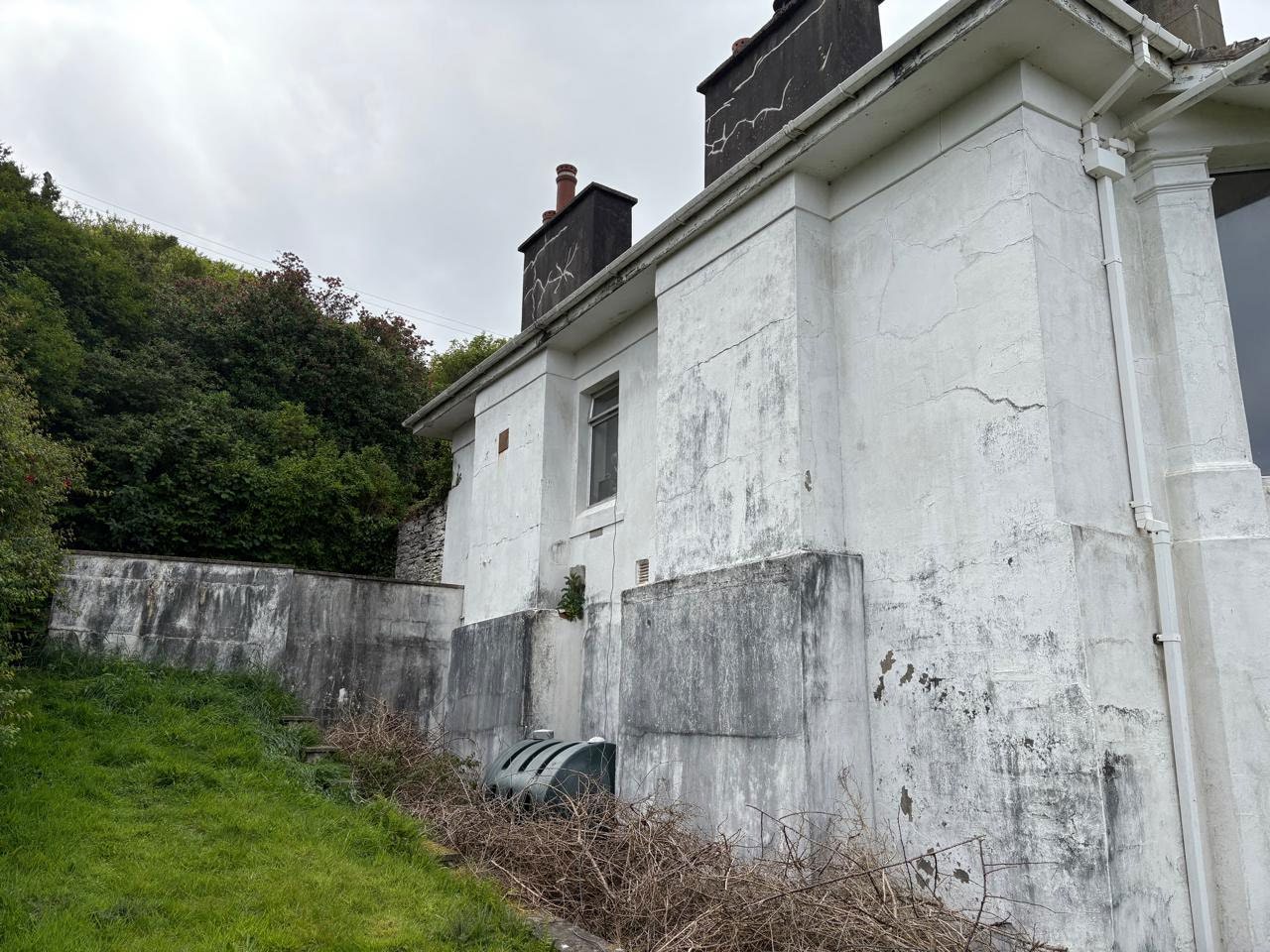 A low-angle photograph showing the weathered white exterior wall of a building with chimneys and guttering, situated next to a grassy slope and retaining wall.
