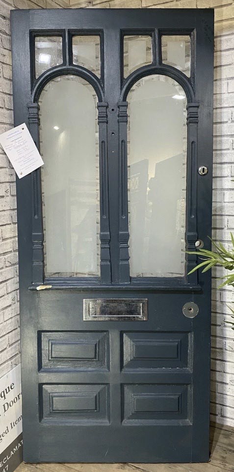 A photograph of a dark blue wooden exterior door with arched glass panels and a letterbox, leaning against a white brick wall.