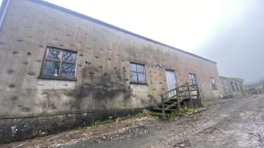 A photograph showing the exterior of a long, single-story concrete outbuilding with a rough, pockmarked facade and wooden steps leading to a door.