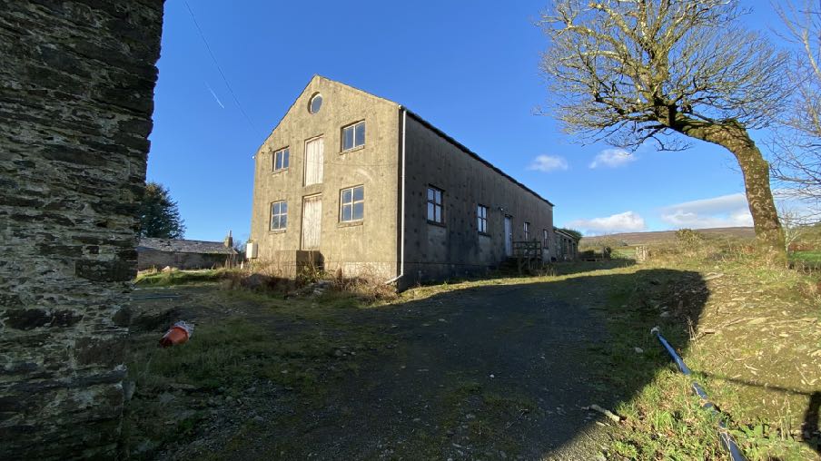 A photograph showing a large, two-story concrete outbuilding with a gable end, situated in a rural setting with a gravel driveway leading up to it.