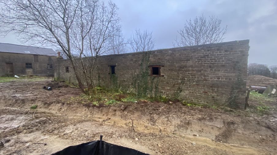 A photograph showing a long, single-story brick outbuilding with open apertures, situated on a site with disturbed earth and a new access path.