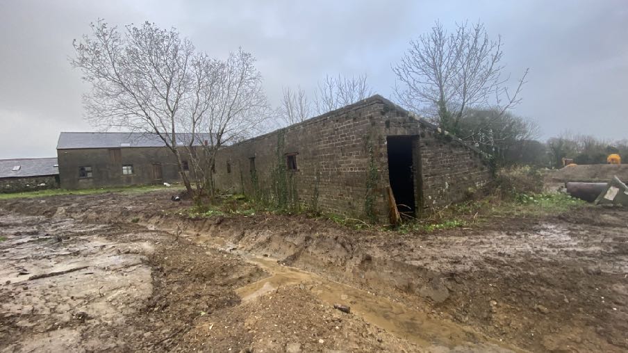 A photograph showing a brick outbuilding situated in a muddy field with visible excavation work in the foreground and another building in the background.