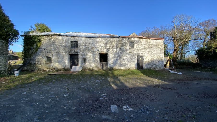 A photograph showing a large, weathered stone outbuilding with a corrugated roof and dark openings, set in a gravel yard.