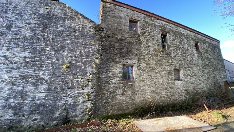 Exterior photograph of a large, weathered stone outbuilding or barn with multiple windows and a pitched roof, showing signs of age and disrepair.