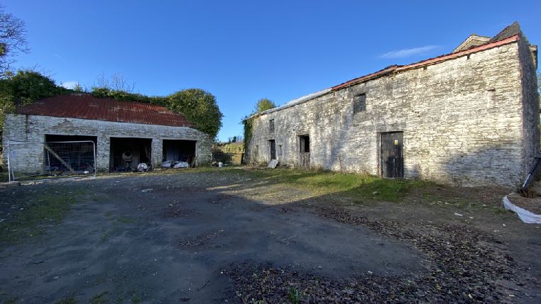 A photograph showing a rural site with two stone outbuildings and a gravel yard. The larger building on the right appears to be a barn, while the smaller structure on the left has open bays.
