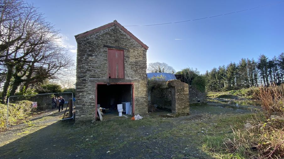 A photograph showing a stone outbuilding or barn with a large ground-floor opening and a smaller upper door, situated in a rural setting with people standing near a temporary fence.