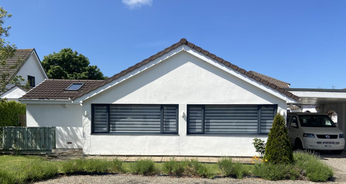 A photograph showing the front elevation of a white single-story bungalow with a gable roof and large louvered windows. A white van is parked to the right under a carport structure.