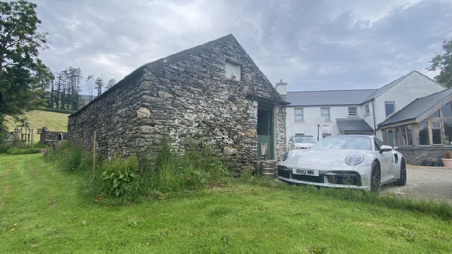 A photograph showing an old stone barn structure adjacent to a modern white house with a driveway and parked car.