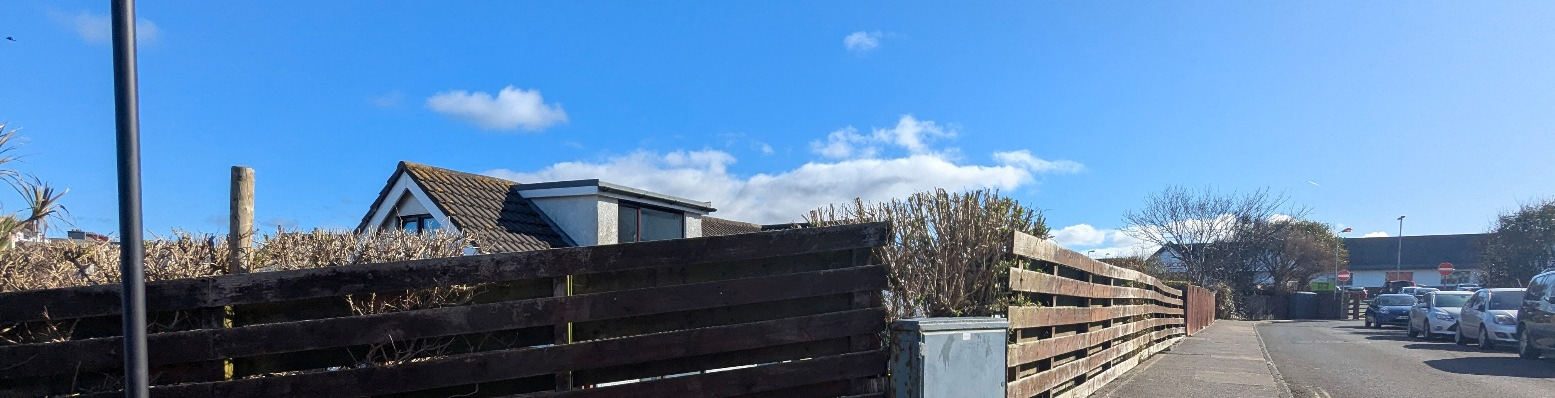 A street-level photograph showing a wooden fence and hedging along a residential property boundary with parked cars visible on the adjacent road.