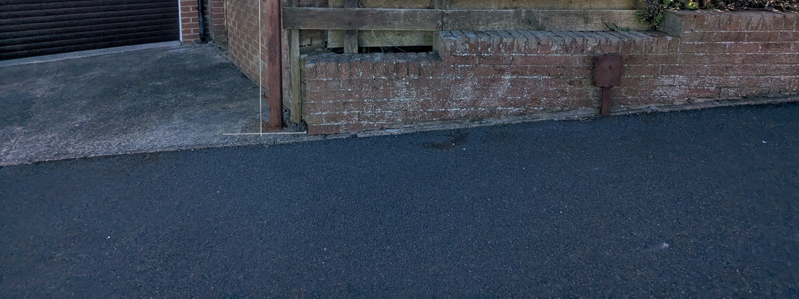 A street-level photograph showing a paved driveway leading to a garage and a low brick wall topped with a wooden fence along the roadside.
