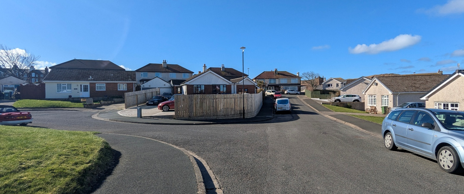 A panoramic photograph of a residential street showing detached houses, a wooden fence, and cars parked on the road and in driveways.
