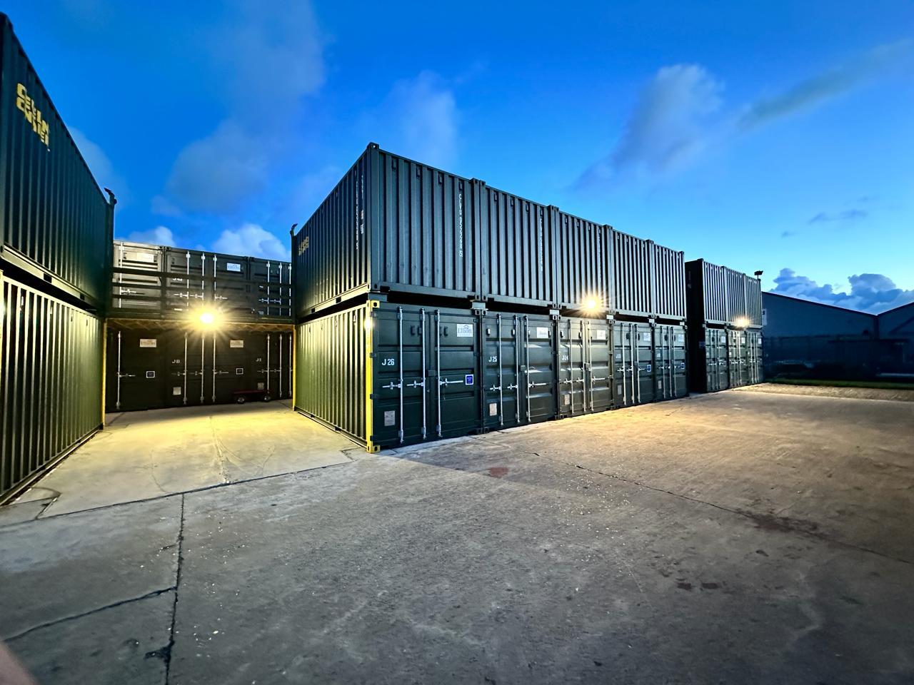 A photograph showing rows of stacked shipping containers used for self-storage, illuminated by artificial lights at dusk.