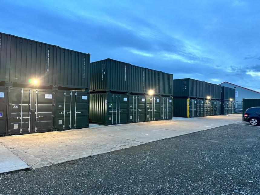 A photograph showing rows of stacked dark shipping containers at a self-storage facility, illuminated by exterior lights against a twilight sky.