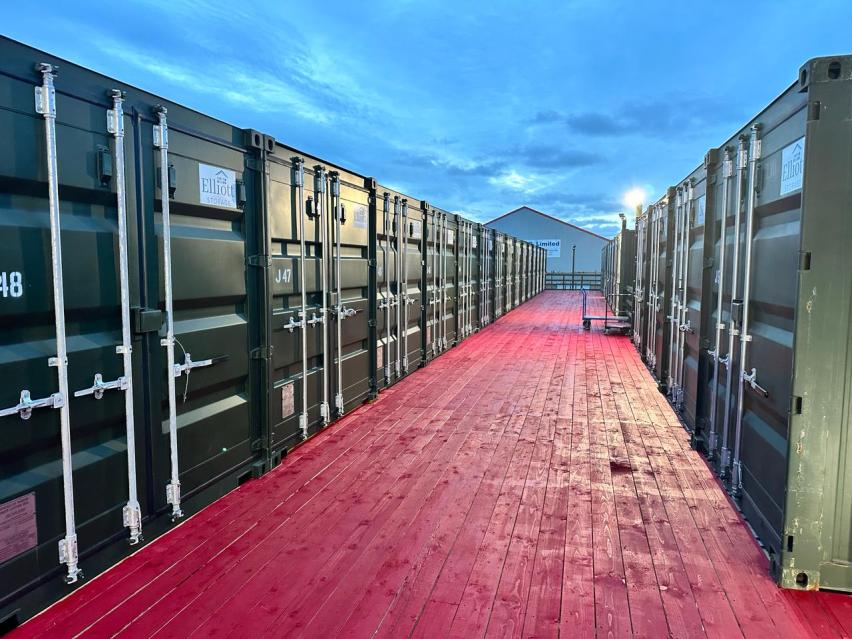 A perspective view of rows of dark green shipping containers flanking a red wooden walkway at a self-storage facility.