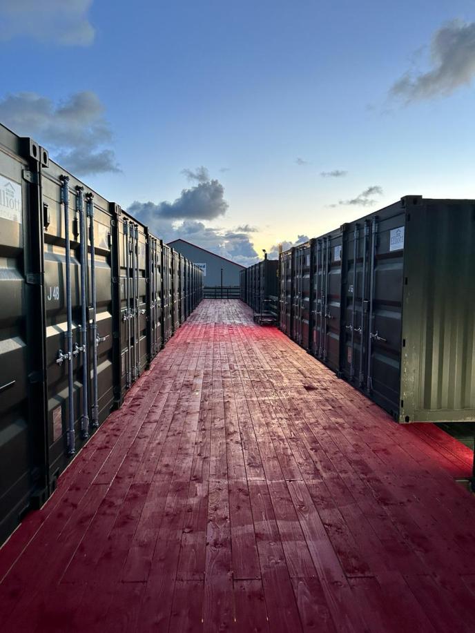 A photograph showing a wooden walkway flanked by two rows of stacked shipping containers at dusk.