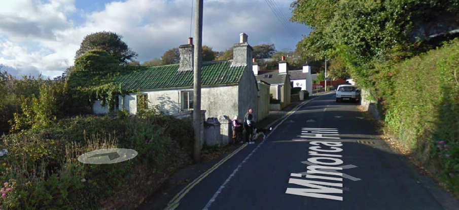 A street-level photograph showing a white cottage with a green roof on the left side of a road labeled 'Minocha Hill', with people standing nearby and a car parked further up the hill.