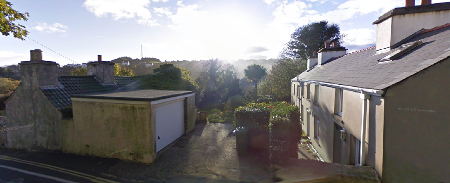 A Google Street View photograph showing existing buildings along a road, including a stone structure with a garage on the left and terraced houses on the right.