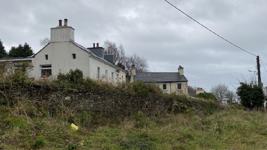 A photograph showing a white detached house and a smaller building behind a stone wall in a rural setting.