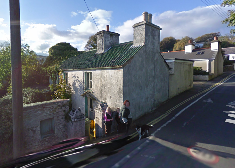 A street-level photograph showing a traditional stone cottage with a slate roof and chimneys situated next to a road with double yellow lines.