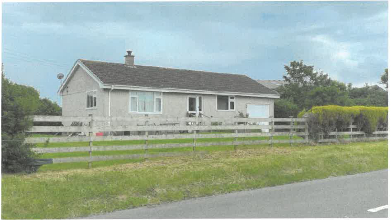 A photograph showing a single-story detached bungalow with a grey tiled roof and white window frames. A wooden post-and-rail fence runs across the front of the property in a rural setting.