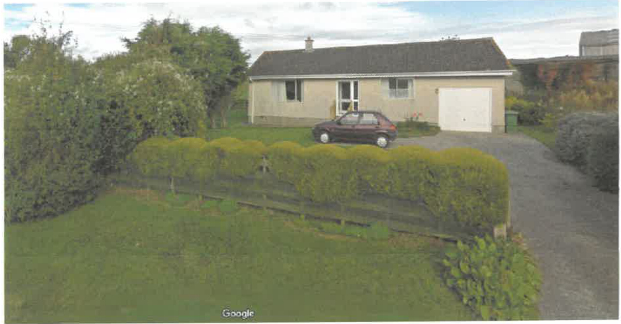 A photograph showing a detached single-story bungalow with an attached garage and a gravel driveway in a rural setting.