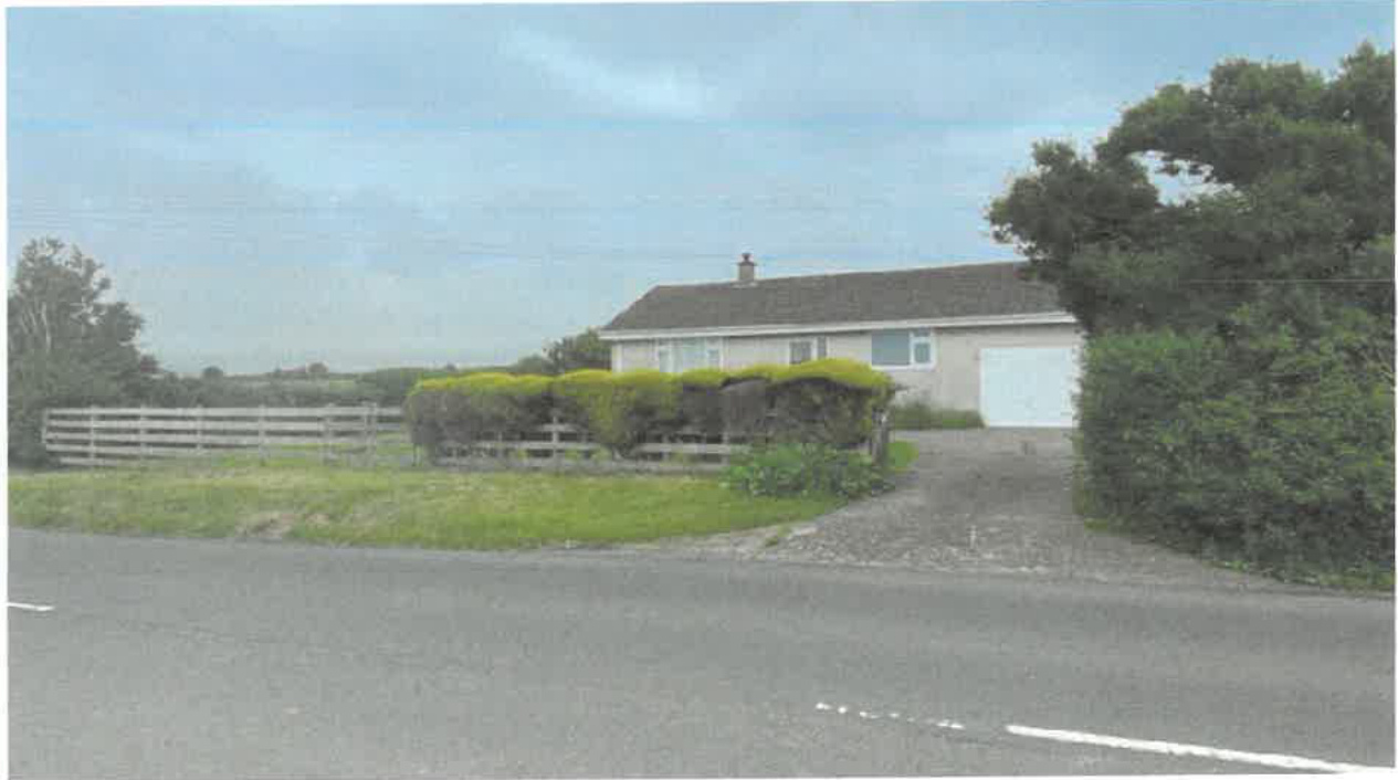 A photograph showing a single-story detached bungalow with an attached garage, situated in a rural setting with a wooden fence and hedges.