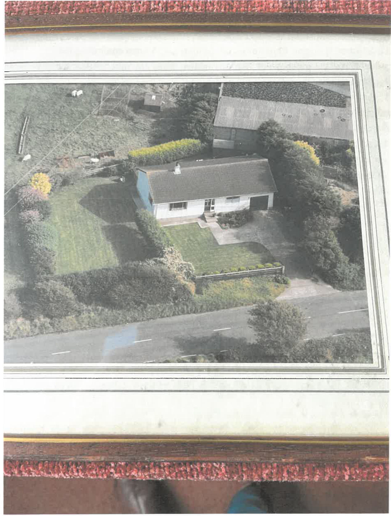 A framed aerial photograph showing a single-story bungalow with a driveway and garden, situated next to a road and an outbuilding.